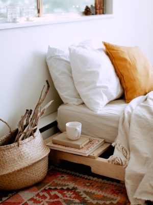 Stylish cozy bedroom interior with bed located near window with white linen, cushions, tray with cup of coffee, basket with dry wooden branches and carpet on floor
