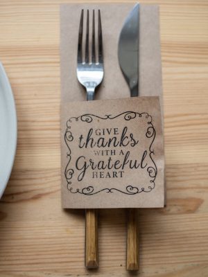 Top view of metal fork and knife in paper cover decorated with Give Thanks With A Grateful Heart inscription placed on wooden table